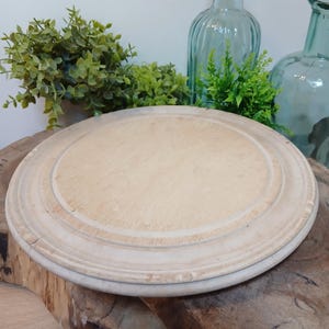 May include: A round, light-coloured wooden serving board with a smooth, flat centre and a raised, grooved circular border. The wood grain is visible across the surface, showing natural variations in tone and texture throughout the circular piece.