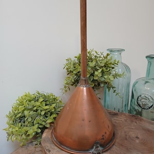 Könnte beinhalten: Ein Kupfertrichter mit langem Holzgriff. Der Trichter hat eine konische Form und einen kleinen Ring an der Seite. Der Hintergrund umfasst grüne Pflanzen und Glasflaschen, was auf eine Vintage- oder rustikale Ästhetik hindeutet.