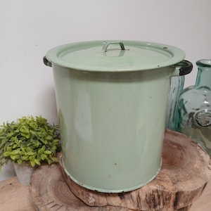May include: A cylindrical light green enamel canister with a matching lid and two dark-rimmed side handles. The metal container features a smooth finish with subtle signs of wear along the edges and a small metal handle on the centre of the lid.