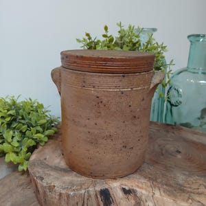 May include: A brown ceramic jar with a lid, featuring two small handles on the sides. The jar has a rustic, speckled appearance and sits on a wooden surface. The lid is a darker shade of brown with horizontal ridges.
