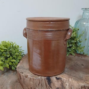 May include: A brown ceramic crock with a lid and two side handles. The cylindrical crock has a textured, ribbed design around the top. It is displayed on a wooden surface, with greenery and a blue glass vase in the background.