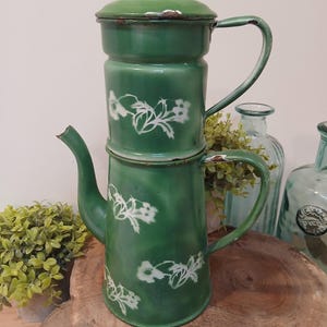 Könnte beinhalten: Vintage grüne Emaille-Kaffeekanne mit weißen Blumenmustern. Die Kanne hat einen Deckel und einen Griff und ist auf einem zweiten Teil gestapelt. Die Kanne steht auf einer Holzoberfläche.