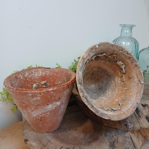 May include: Two weathered terracotta pots with a conical shape and wide, open rims. The exterior surfaces show a rustic, reddish-brown finish with visible signs of wear, mineral deposits, and textured patches along the edges and inner walls.