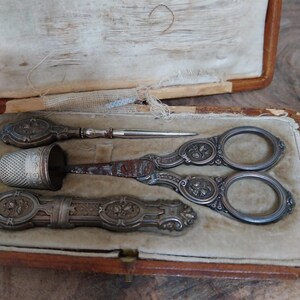 May include: An antique sewing kit featuring ornate silver scissors, a thimble, and a needle holder. The items are presented in a worn, open, brown leather case with a cream interior.