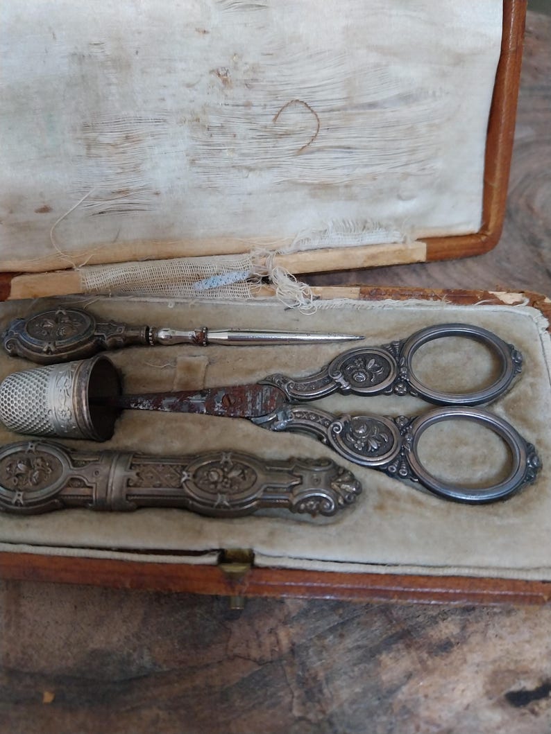 May include: An antique sewing kit featuring ornate silver scissors, a thimble, and a needle holder. The items are presented in a worn, open, brown leather case with a cream interior.