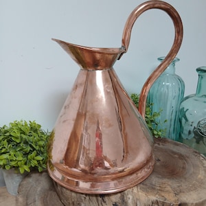 May include: A polished copper pitcher with a wide, conical base, a narrow neck, and a tall, curved handle. The metal surface is reflective, showing light and surrounding shapes. The pitcher features a flared spout for pouring liquids.