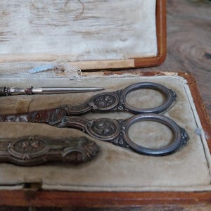 May include: An antique sewing kit in a brown leather case. The kit includes ornate silver scissors, a needle, and a small tool, all on a beige velvet lining. The scissors and tools feature floral designs.