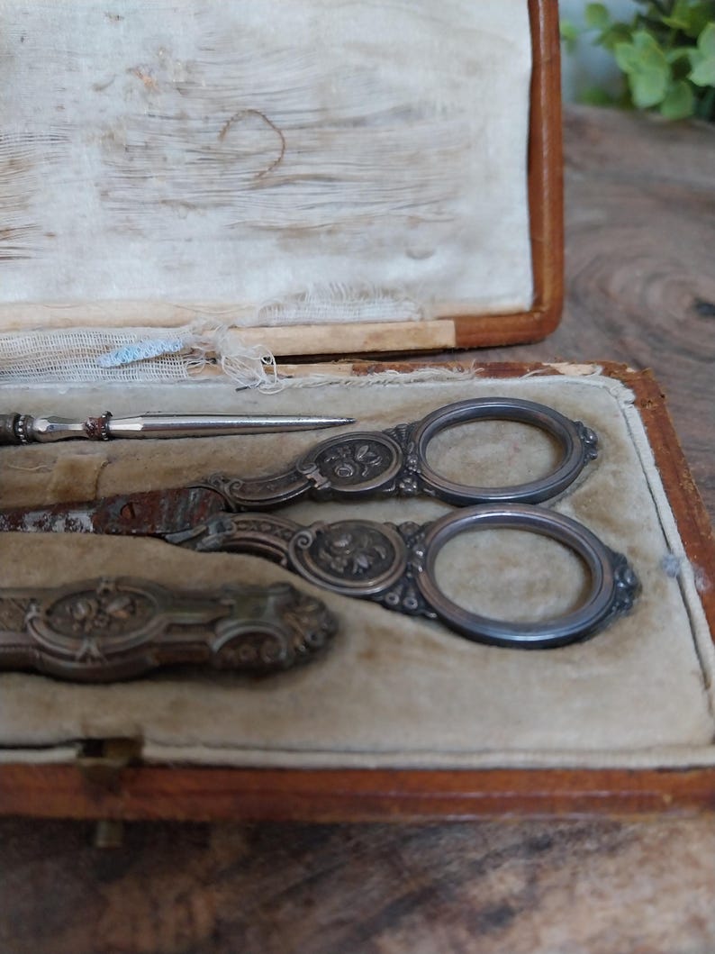 May include: An antique sewing kit in a brown leather case. The kit includes ornate silver scissors, a needle, and a small tool, all on a beige velvet lining. The scissors and tools feature floral designs.