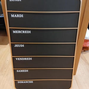 May include: A rectangular black weekly planner with gold trim and horizontal gold accents. The days of the week are printed in white: LUNDI, MARDI, MERCREDI, JEUDI, VENDREDI, SAMEDI, and DIMANCHE. The planner is hanging on a wall.