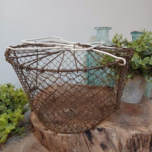 Op de afbeelding: Een vintage draadmand met een roestbruine afwerking en een witte handgreep. De mand heeft een afgeronde vorm en een gaasontwerp. Hij staat op een houten oppervlak, met groen en blauwe glazen flessen op de achtergrond. Perfect voor landelijke decoratie.
