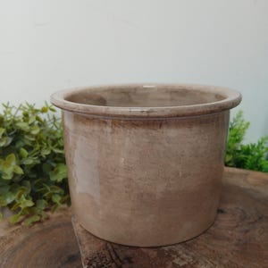 May include: A cylindrical ceramic crock featuring a smooth, light beige finish with subtle brown mottled variations throughout the surface. The vessel has a wide, rounded rim and a flat base, presenting a simple, functional design for storage.