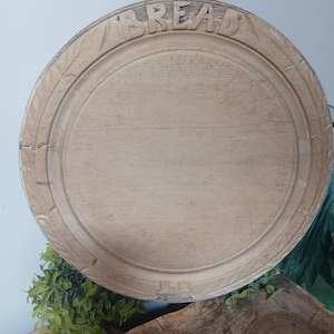 Puede incluir: Una tabla de pan redonda de madera clara con un borde tallado con patrones en forma de hoja. La palabra BREAD está grabada en letras mayúsculas a lo largo del borde superior y la superficie muestra la veta natural de la madera.