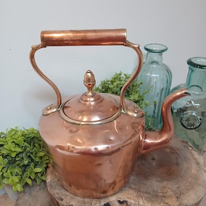 Puede incluir: Una tetera de cobre con un pico curvo y un asa de madera. La tetera tiene una tapa con un pomo decorativo. Es de color marrón rojizo y está colocada sobre una superficie de madera.