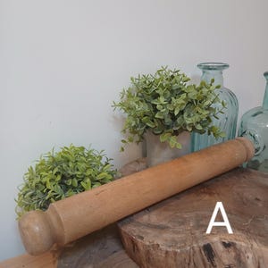 Può includere: Un mattarello in legno marrone chiaro con estremità arrotondate, appoggiato su un tagliere di legno. Lo sfondo presenta piante verdi in vaso e due bottiglie di vetro blu. Un utensile da cucina.
