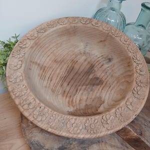 May include: A round wooden bowl featuring a carved floral pattern along the wide rim. The bowl displays a natural light brown wood grain with visible rings and texture throughout the interior surface and the carved flower details on the edge.