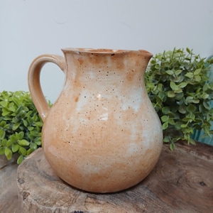 May include: A handmade ceramic jug with a rounded body and a curved handle. The jug has a speckled, light orange and white glaze. The rim and handle are a darker orange. The jug is sitting on a wooden surface.