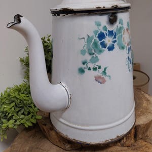 Puede incluir: Cafetera de esmalte blanco con diseño floral en azul, verde y rosa. La cafetera vintage tiene un pico curvo, una tapa con un pequeño pomo y muestra signos de desgaste. Se encuentra sobre una superficie de madera, sugiriendo una estética rústica.