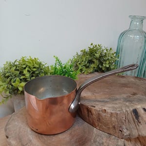 Puede incluir: Un cazo de cobre con un exterior metálico pulido y un mango largo y curvado de metal plateado. El interior del cazo es de color plateado y presenta un pequeño pico vertedor en el borde, con un diseño cilíndrico clásico.