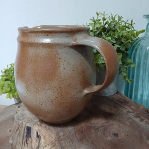 Può includere: Una tazza in ceramica fatta a mano con un corpo arrotondato e un manico grande. La tazza presenta una smaltatura marrone terrosa macchiettata, con toni più chiari vicino al bordo e al manico. Appoggiata su una superficie di legno.