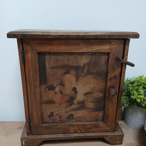 May include: A rectangular wooden cabinet with a single hinged door featuring a printed image of chickens, a rooster, and chicks in a farm setting. The cabinet has a dark brown wood finish, a small round wooden knob, and a latch on the right side.