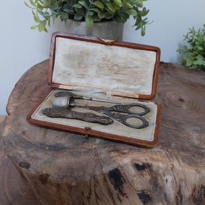 May include: A vintage sewing kit presented in a brown leather case. The kit contains a thimble, scissors, and a needle holder, each with intricate silver-toned embellishments. The case is open, displaying the tools set within a cream-coloured fabric lining.