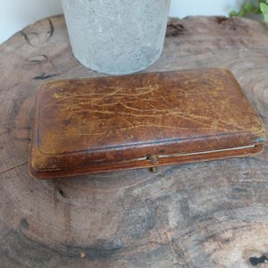 May include: A rectangular, antique brown leather box with a clasp. The box has a worn, textured surface with visible scratches and age marks. The box is closed and appears to be empty. The box is sitting on a wooden surface.