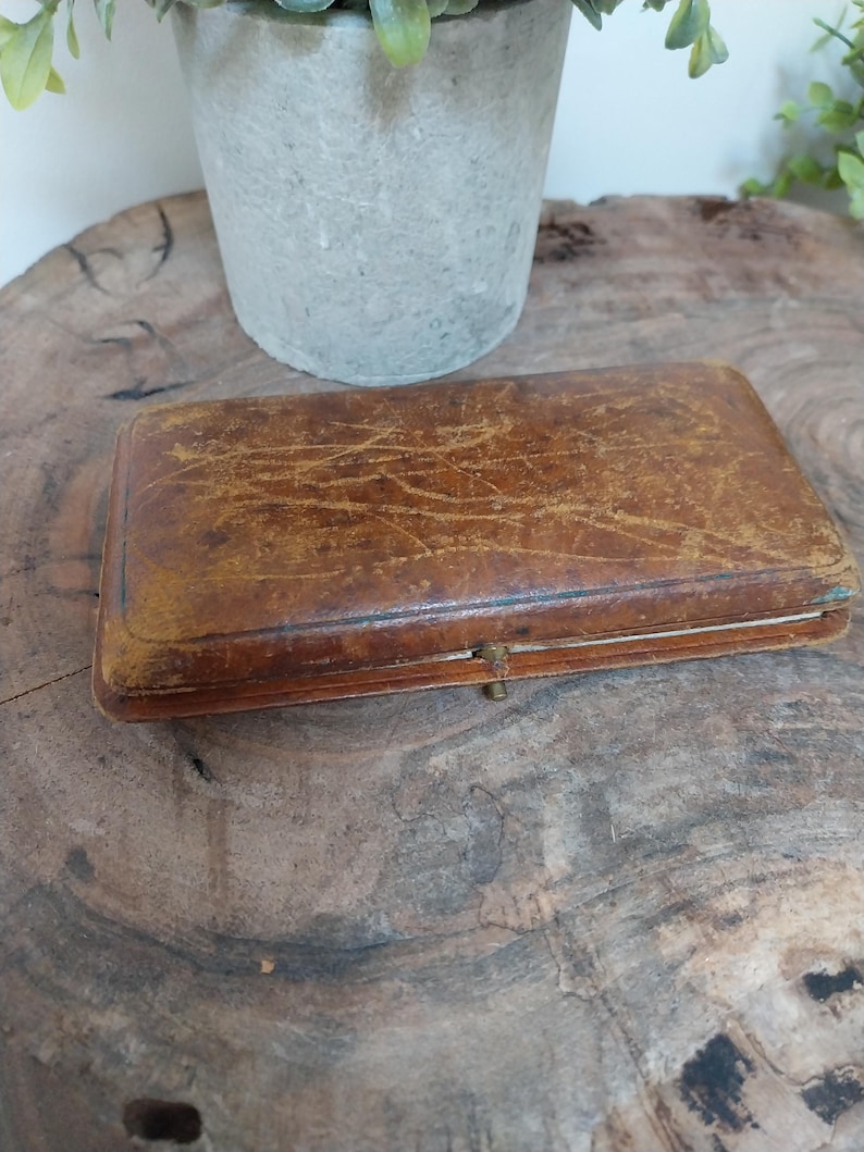 May include: A rectangular, antique brown leather box with a clasp. The box has a worn, textured surface with visible scratches and age marks. The box is closed and appears to be empty. The box is sitting on a wooden surface.