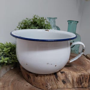 Peut inclure: Un pot de chambre en émail blanc avec un bord et une poignée bleus. Le pot présente des signes d'usure, ce qui ajoute à son charme vintage. Il est posé sur une surface en bois, avec du feuillage vert et des bouteilles en verre bleues en arrière-plan.