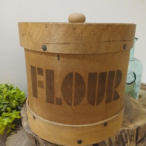以下が含まれることがあります： ふた付きのヴィンテージ木製小麦粉容器。側面には「FLOUR」の文字が大きく、濃い色でステンシルされています。容器は円筒形で、明るい茶色の仕上げで、小さな金属リベットで固定されています。