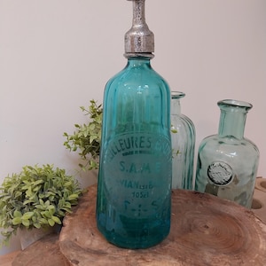 May include: A teal glass bottle with a silver metal stopper. The bottle has embossed text and is 10.5 cl. Other glass bottles are in the background. The bottle is displayed on a wooden surface.