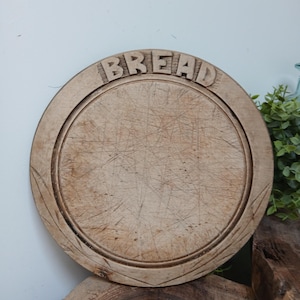 Pode incluir: Uma tábua de pão redonda de madeira com a palavra BREAD esculpida na borda superior. A superfície apresenta marcas de faca e arranhões visíveis devido ao uso, com um sulco circular ao longo do perímetro interno da madeira clara.