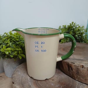 May include: A cream-coloured enamel measuring jug with a green rim and handle. The exterior features blue printed text reading OZ. 20, PT. 1, and GR. 500. The interior rim displays measurement markings for OZ, PT, and GR with a vertical line.