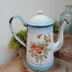 Könnte beinhalten: Eine hohe, zylindrische Emaille-Kaffeekanne mit weißem Körper und hellblauem Farbverlauf an Deckel und Boden. Die Außenseite ist mit einem Blumenmotiv aus Stiefmütterchen in Orange-, Gelb- und Grüntönen verziert, mit sichtbarem Rost an den Rändern.