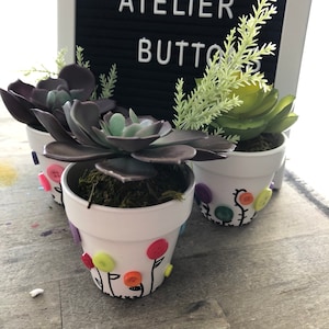 May include: Three small white flower pots with green succulents and decorated with colorful buttons. The pots are labeled "Cactus Atelier Buttons".