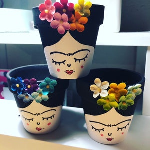 May include: Three black terracotta flower pots with painted faces and colorful flower accents. The faces are painted with closed eyes, a nose, and a red mouth. The flowers are painted in shades of pink, red, yellow, blue, and green.