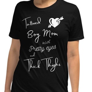 May include: Black t-shirt with white text that reads "Tattooed Boy Mom with Pretty Eyes and Thick Thighs". The text includes a heart with an arrow through it.