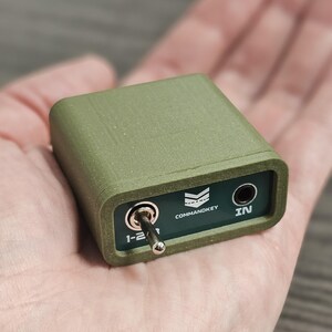 Op de afbeelding: Een olijfgroen elektronisch apparaat met een tuimelschakelaar en een 6,35 mm audio-aansluiting. Het apparaat heeft de tekst "COMMANDKEY" en "IN" erop gedrukt. Het apparaat wordt in een hand gehouden.