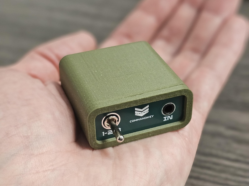 Op de afbeelding: Een olijfgroen elektronisch apparaat met een tuimelschakelaar en een 6,35 mm audio-aansluiting. Het apparaat heeft de tekst "COMMANDKEY" en "IN" erop gedrukt. Het apparaat wordt in een hand gehouden.