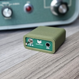 Op de afbeelding: Een klein, olijfgroen elektronisch apparaat met een groen frontpaneel. Op het frontpaneel staat de tekst "COMMANDKEY" en "IN". Een tuimelschakelaar staat in de stand "1-2-3". Het apparaat is rechthoekig en heeft afgeronde randen.