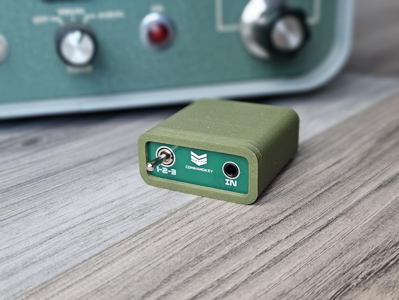 Op de afbeelding: Een klein, olijfgroen elektronisch apparaat met een groen frontpaneel. Op het frontpaneel staat de tekst "COMMANDKEY" en "IN". Een tuimelschakelaar staat in de stand "1-2-3". Het apparaat is rechthoekig en heeft afgeronde randen.