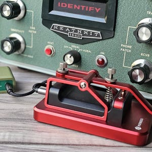 Op de afbeelding: Een vintage Heathkit SB-630 stationconsole met een rode morsecode-sleutel en een kleine groene doos. De console is olijfgroen met diverse knoppen en de tekst "IDENTIFY" en "HEATHKIT SB-630". De sleutel is verbonden met de groene doos.