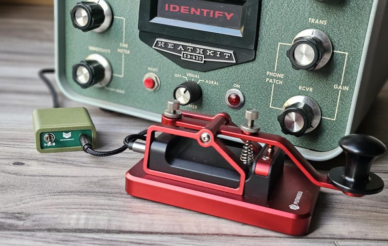 Op de afbeelding: Een vintage Heathkit SB-630 stationconsole met een rode morsecode-sleutel en een kleine groene doos. De console is olijfgroen met diverse knoppen en de tekst "IDENTIFY" en "HEATHKIT SB-630". De sleutel is verbonden met de groene doos.