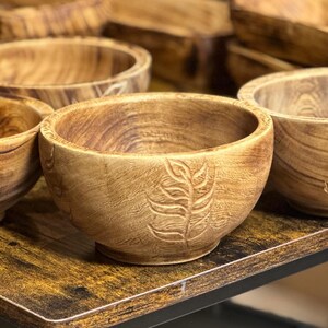 Puede incluir: Un conjunto de cinco cuencos de madera con un dise&ntilde;o de hojas tallado en la superficie. Los cuencos est&aacute;n dispuestos sobre una mesa de madera.