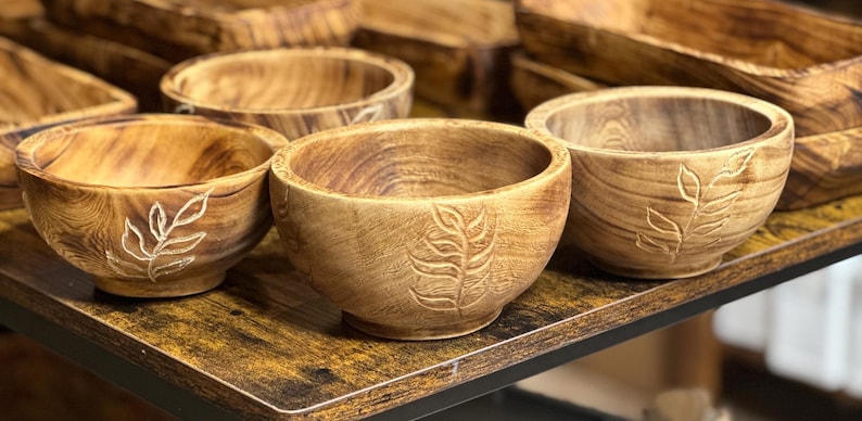 Puede incluir: Un conjunto de cinco cuencos de madera con un dise&ntilde;o de hojas tallado en la superficie. Los cuencos est&aacute;n dispuestos sobre una mesa de madera.