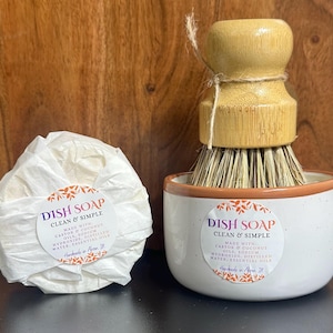 May include: A round bar of dish soap wrapped in white paper with a label reading DISH SOAP CLEAN & SIMPLE, alongside a white ceramic bowl containing a bamboo dish brush with natural bristles and a matching label on the front of the bowl.