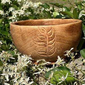 Puede incluir: Un cuenco de madera con un dise&ntilde;o de hoja tallada. El cuenco es de color marr&oacute;n claro y tiene un acabado de madera natural. Est&aacute; sentado en un lecho de flores blancas.
