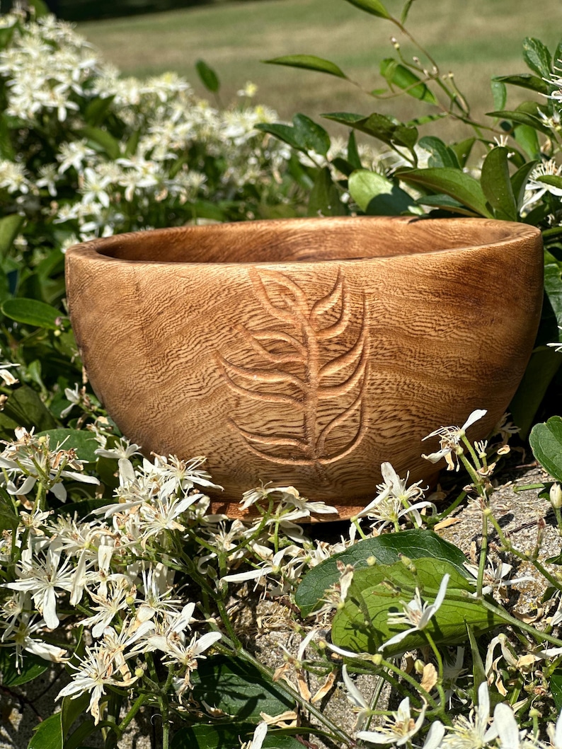 Puede incluir: Un cuenco de madera con un dise&ntilde;o de hoja tallada. El cuenco es de color marr&oacute;n claro y tiene un acabado de madera natural. Est&aacute; sentado en un lecho de flores blancas.