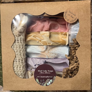 May include: A gift box filled with handmade soaps in a variety of colors and scents. The soaps are stacked on top of each other and are surrounded by brown paper shreds. The box is decorated with a brown paper label that says "Mud Lake Soaps, Akron, Indiana - handcrafted".