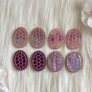 May include: Eight oval-shaped resin cabochons with a gold-toned geometric design. The cabochons have a gradient of purple and pink colors.