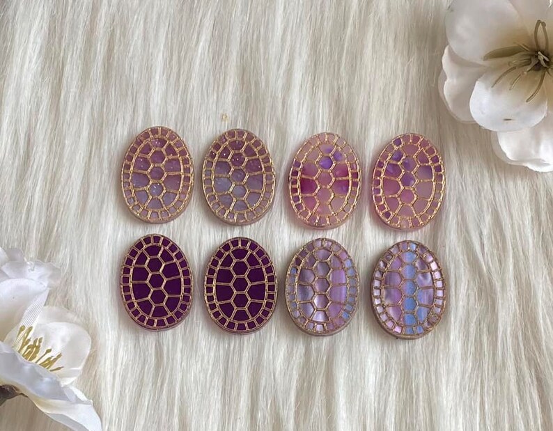 May include: Eight oval-shaped resin cabochons with a gold-toned geometric design. The cabochons have a gradient of purple and pink colors.